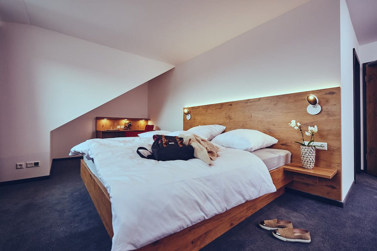 A modern hotel room with a wooden bed, a bedside table with a lamp, a small desk with a chair, and a wall-mounted shelf with a vase of flowers.