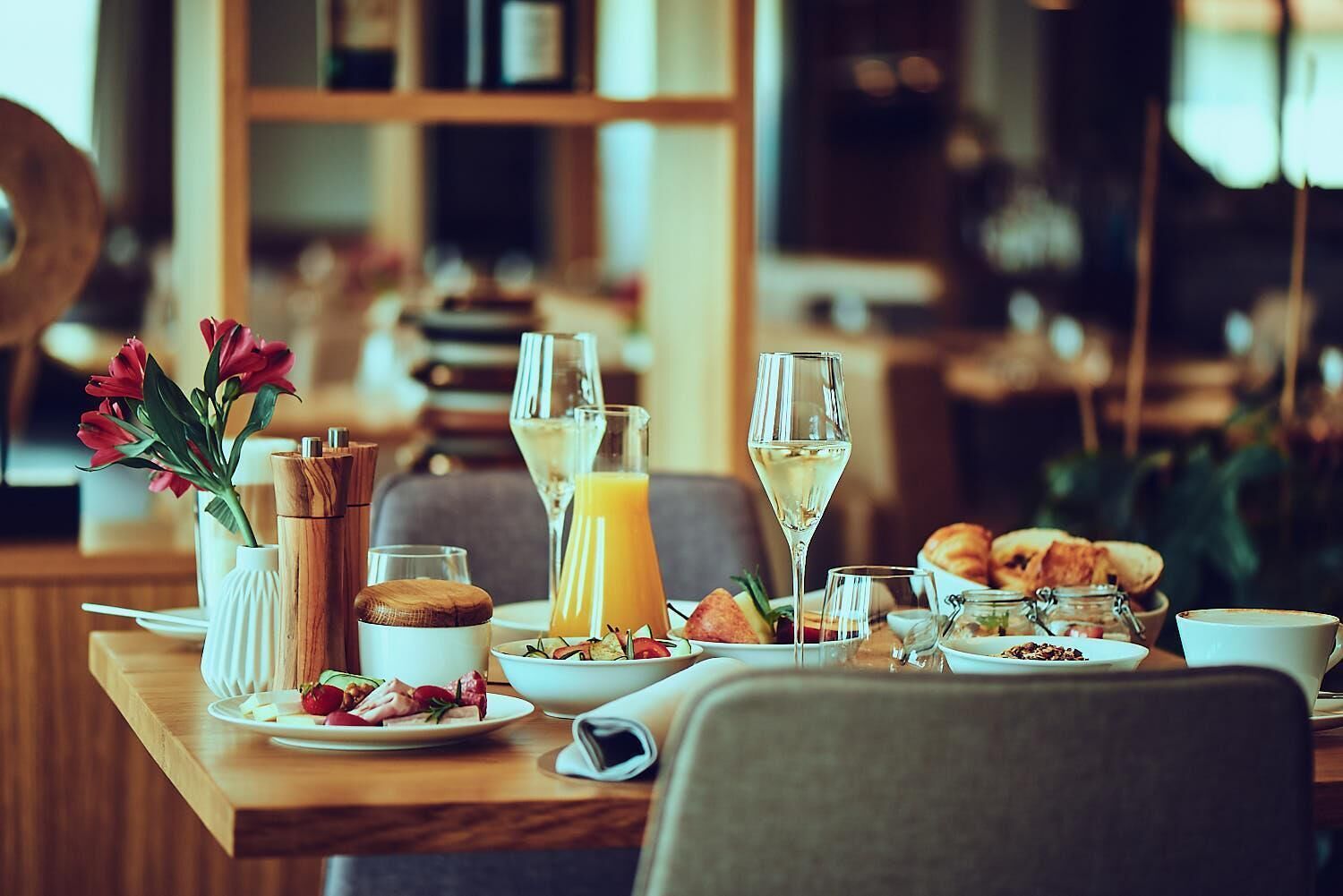 Een ontbijtsetting met een gedekte tafel voor twee personen, inclusief borden met eten, glazen sap en wijn, en een vaas rode bloemen.