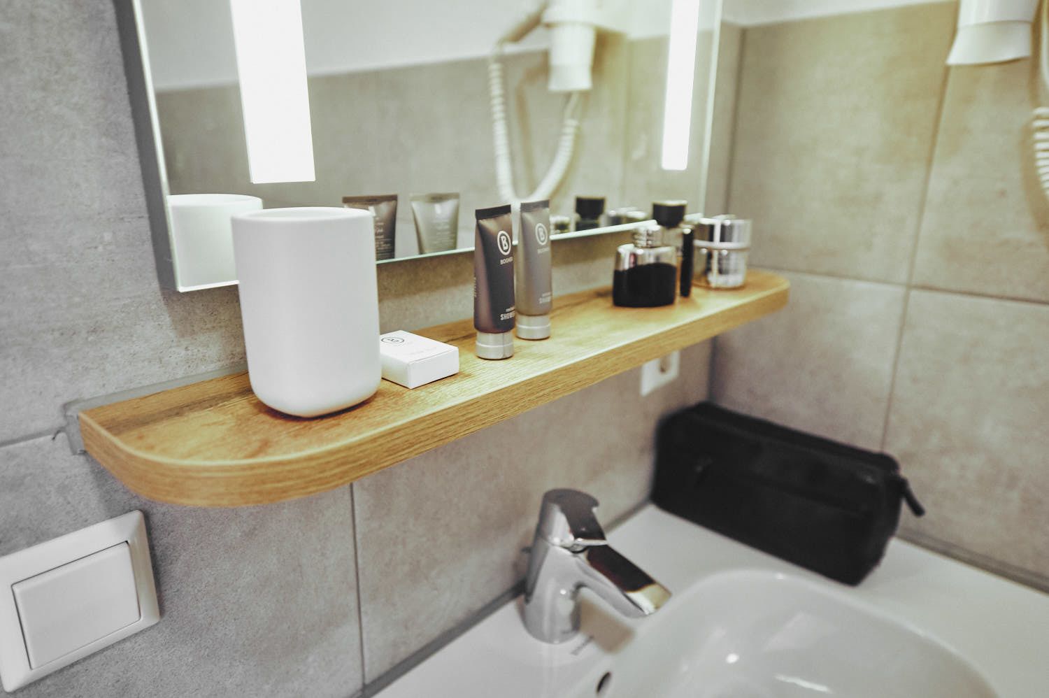 A modern bathroom with a wooden shelf holding toiletries, a hairdryer, and a mirror above a sink.