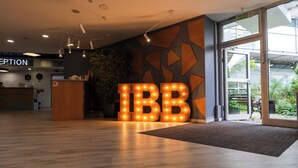 Un área de recepción con un mostrador de madera, un gran letrero iluminado que dice 'IBB' y una puerta de vidrio que da al exterior.