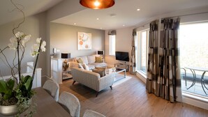 A modern living room with a sofa, coffee table, and TV. There is a large window with curtains, a potted plant, and a painting on the wall. - Beech House (Bristol)
