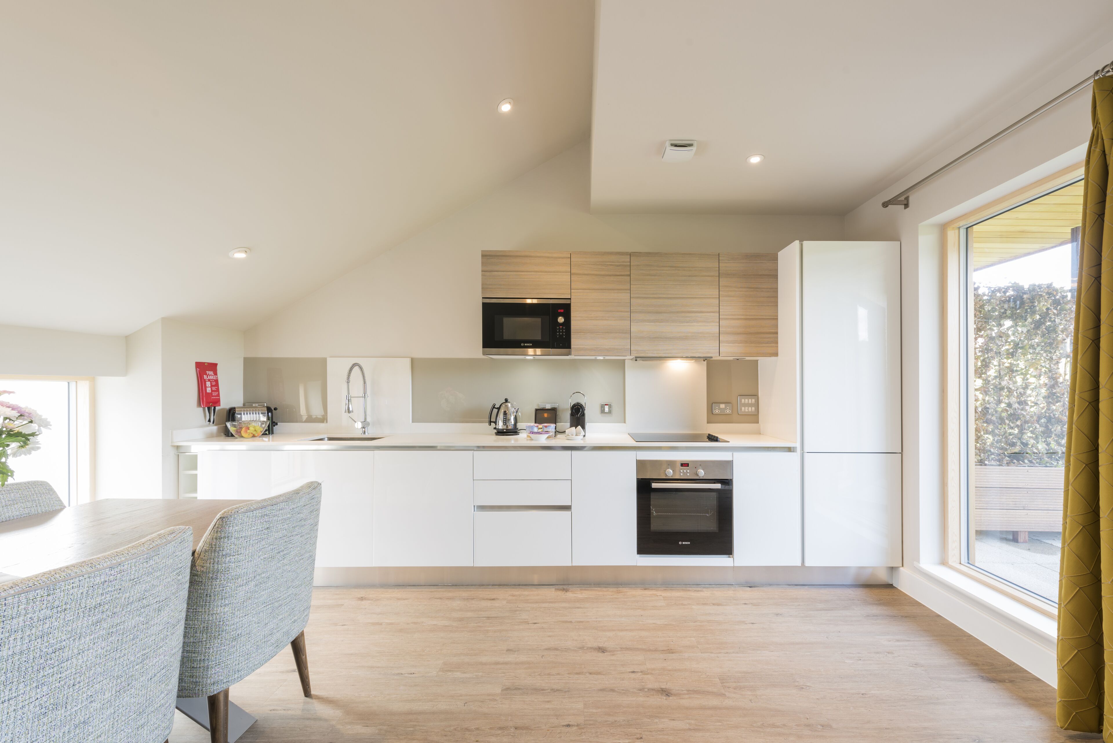 A modern kitchen with white cabinetry, a built-in microwave, and an oven. There is a dining table with chairs and a window providing a view of the outdoors.