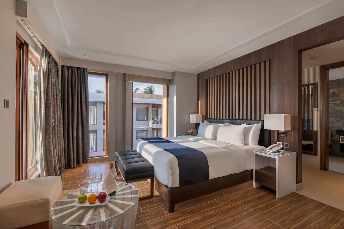 Quarto de hotel moderno com uma cama grande, uma mesa de centro de vidro com frutas, uma pequena mesa lateral com um telefone e vista para a varanda externa.