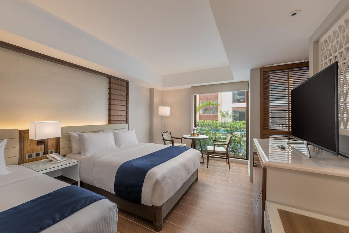 Quarto de hotel com duas camas, uma escrivaninha com luminária, TV e varanda com mesa e cadeiras.