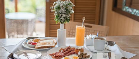 A breakfast tray with toast, eggs, bacon, mushrooms, and a glass of orange juice on a table with a white vase of flowers.