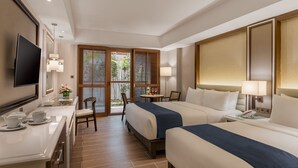 A hotel room with two beds, a desk with a flat-screen TV, a small table with a lamp, and a view of greenery through the window.