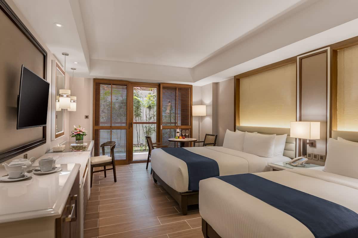 Quarto de hotel com duas camas, uma escrivaninha com televisão de tela plana, uma mesinha com abajur e vista para o verde pela janela.