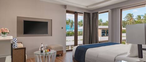 A modern hotel room with a large bed, a flat-screen TV mounted on the wall, a small round table with a glass top, and a view of the beach and palm trees through the windows.