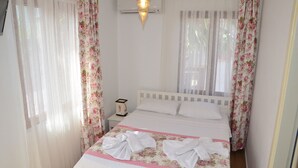 A neatly made bed with floral bedding and towels, a bedside table with a clock, and sheer curtains. - Mercanay Butik Otel (Ayvalik)