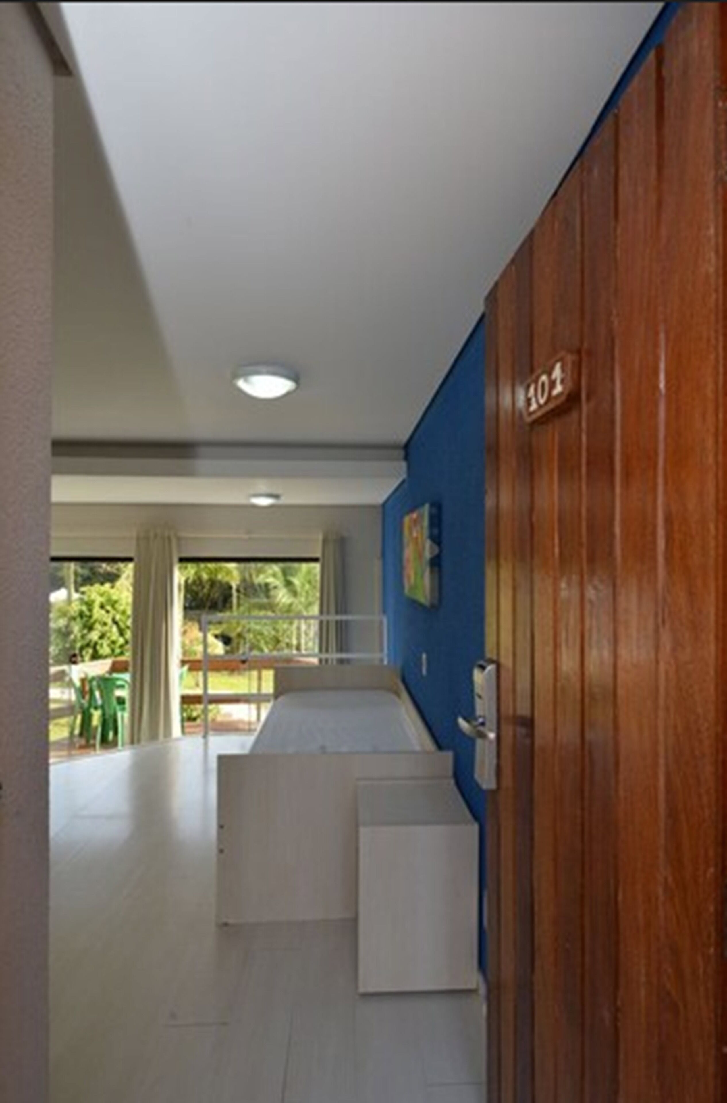 Habitación de hotel con una puerta de madera marcada con el número 101, un escritorio pequeño y vistas al exterior por una ventana.