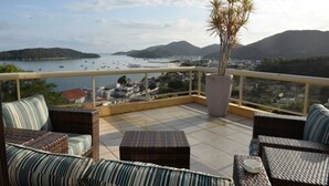 Un balcón con mobiliario de mimbre, una planta en maceta y vista al mar y las colinas.