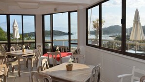 Un restaurante con amplios ventanales que ofrece vistas al mar y a las colinas. Las mesas están dispuestas con sillas, y se observan sombrillas en el exterior.