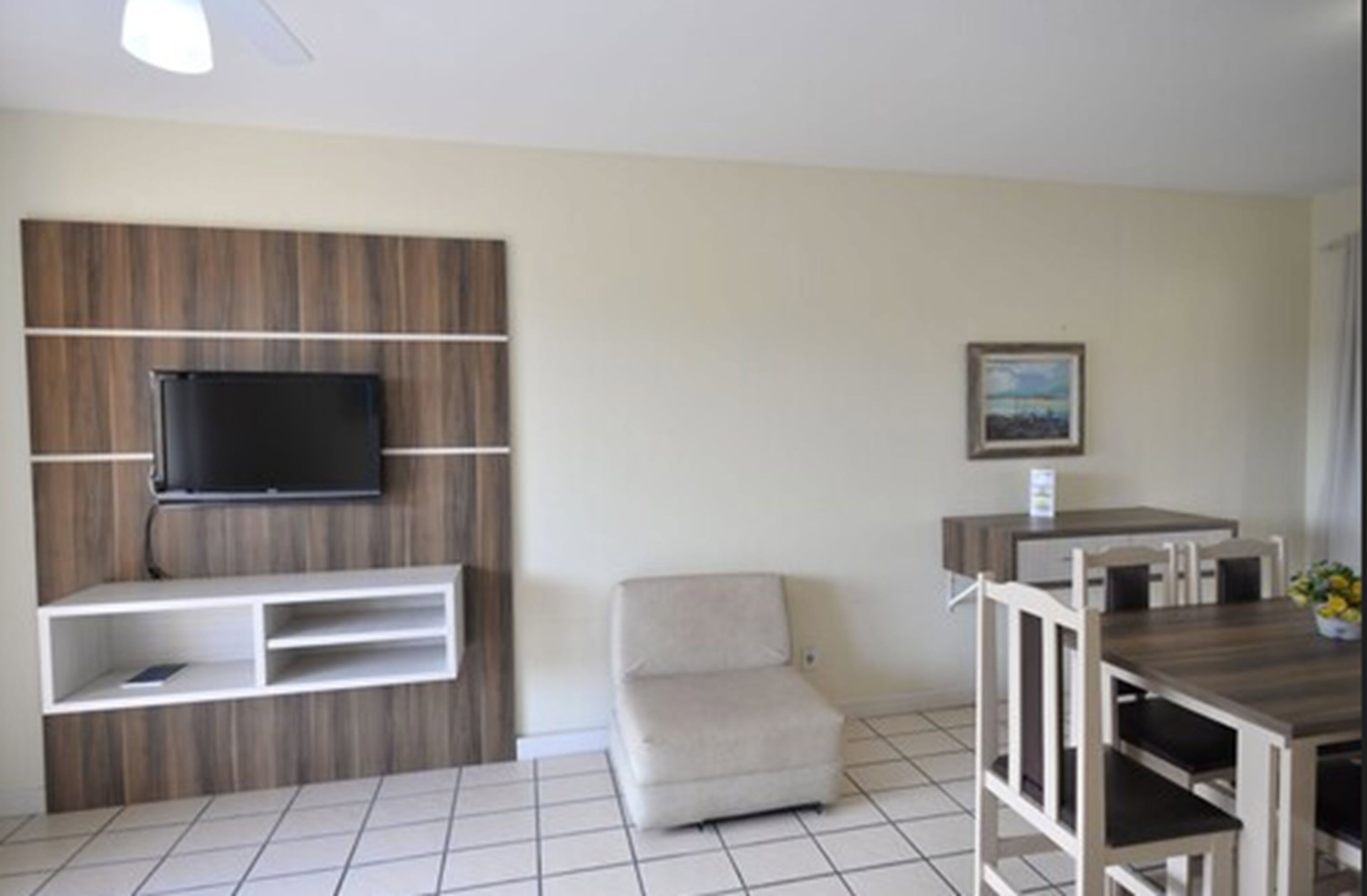 A modern living room with a flat-screen TV mounted on a wooden panel wall, a small beige armchair, a dining table with chairs, and a framed picture on the wall.
