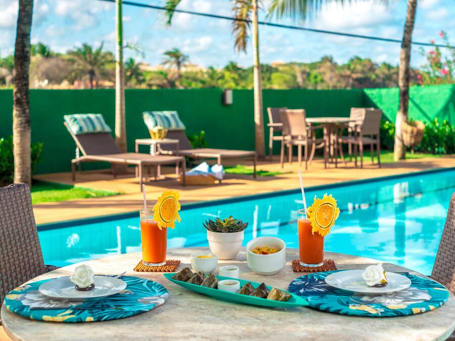 Zona de comedor junto a la piscina, con una mesa lista para dos personas, con temática tropical y una decoración en tonos azules y amarillos.