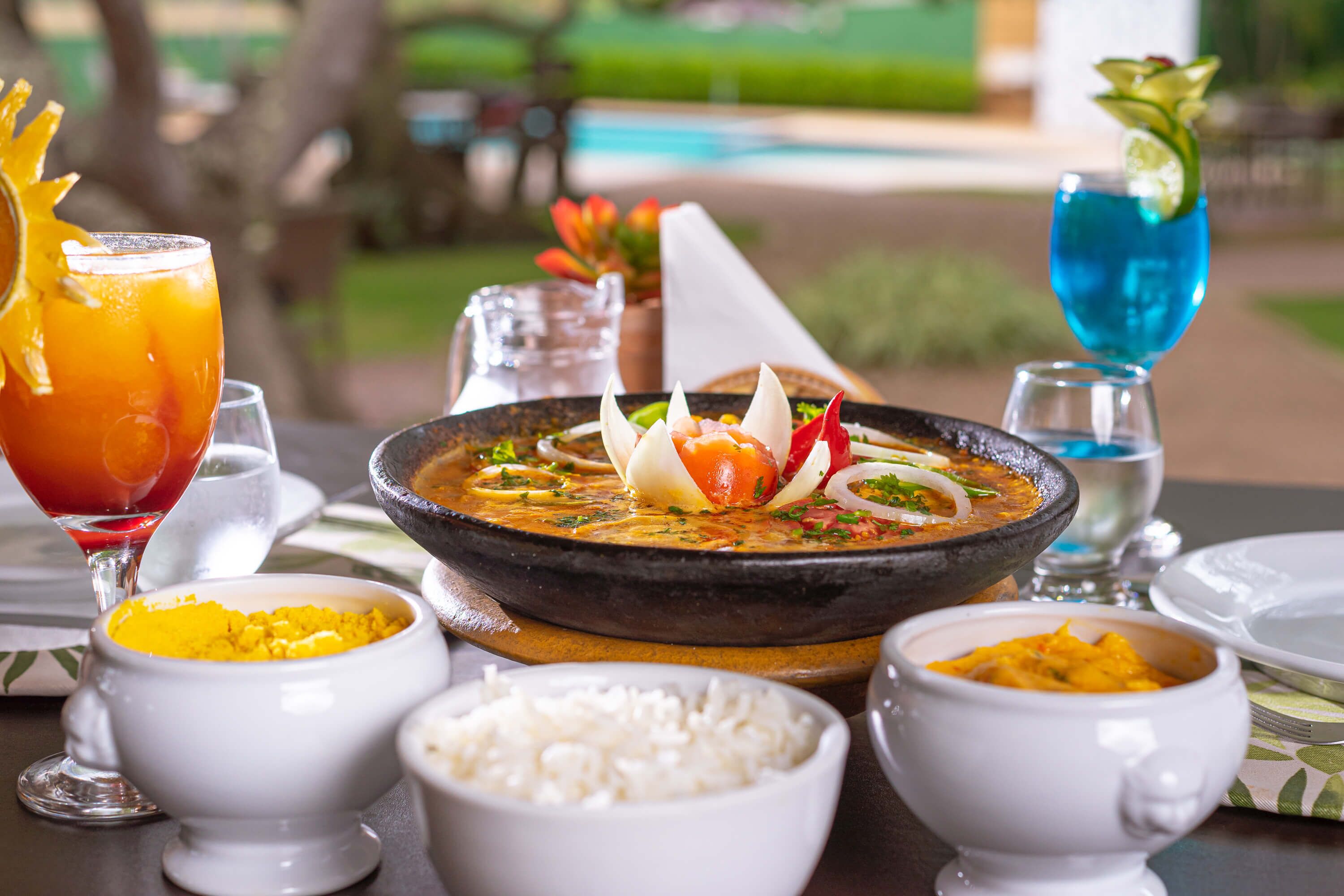 Une table à manger est dressée avec une variété de plats, notamment un bol de riz, un bol de nourriture jaune, un bol de nourriture orange, un bol de nourriture blanche et un bol de nourriture verte. On y trouve également deux boissons, l’une dans un verre et l’autre dans un bol, ainsi qu’un objet décoratif sur la table.