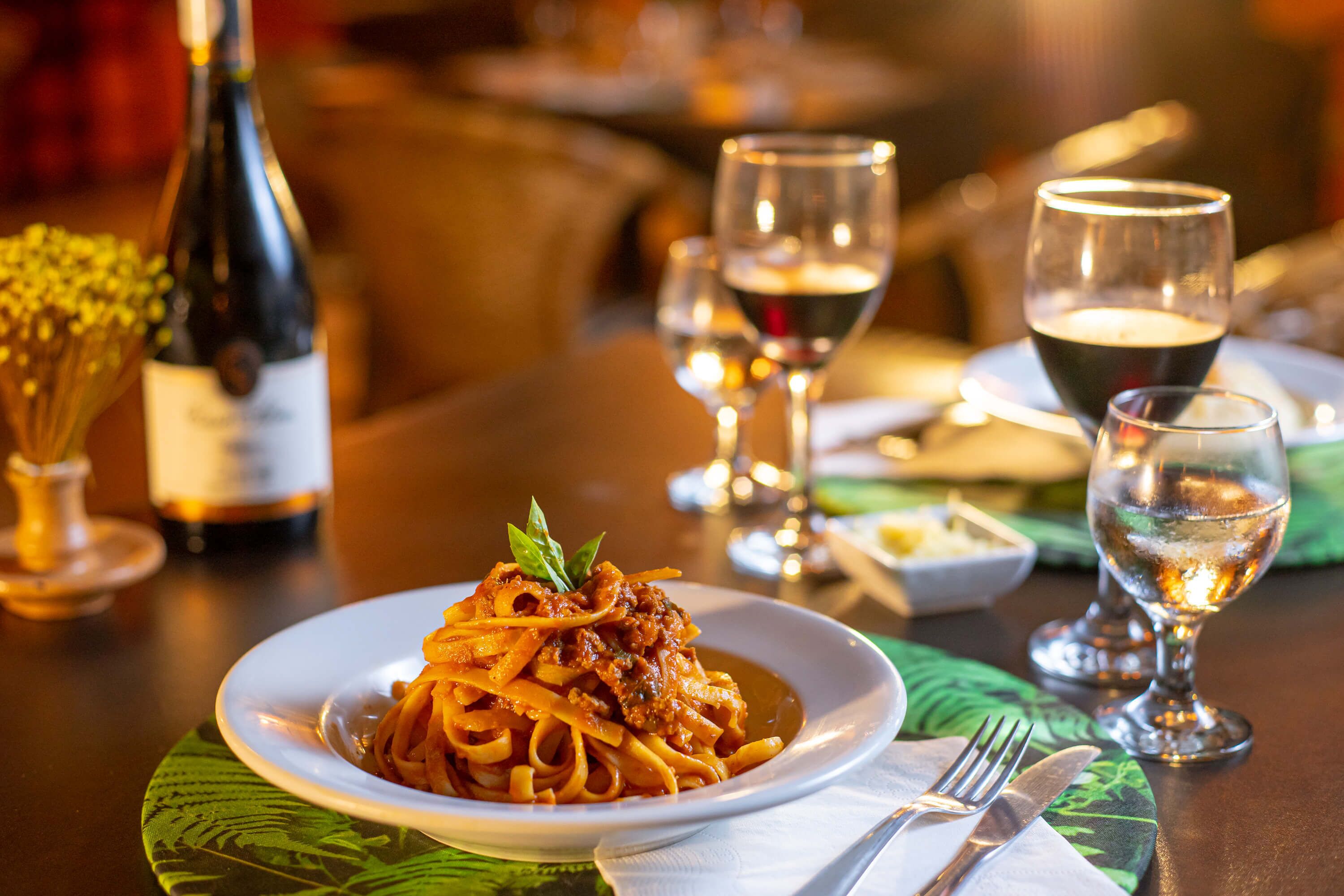 A dining table with a plate of spaghetti, wine glasses, and a bottle of wine.