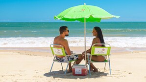 Een stel zit onder een groene parasol op het strand, met een koelbox ernaast.