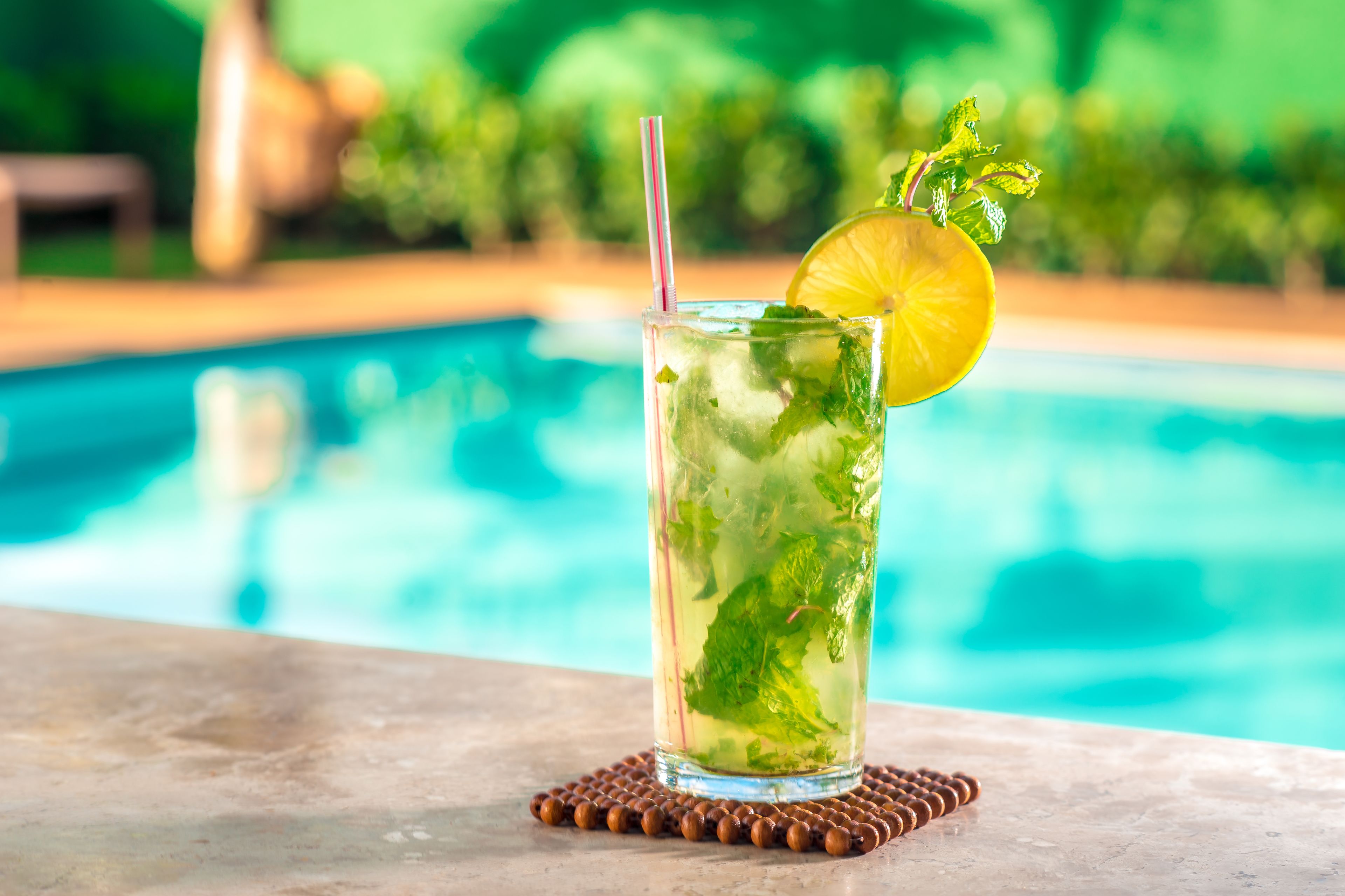 A glass of iced beverage with mint and a slice of lime, placed on a coaster by a pool.