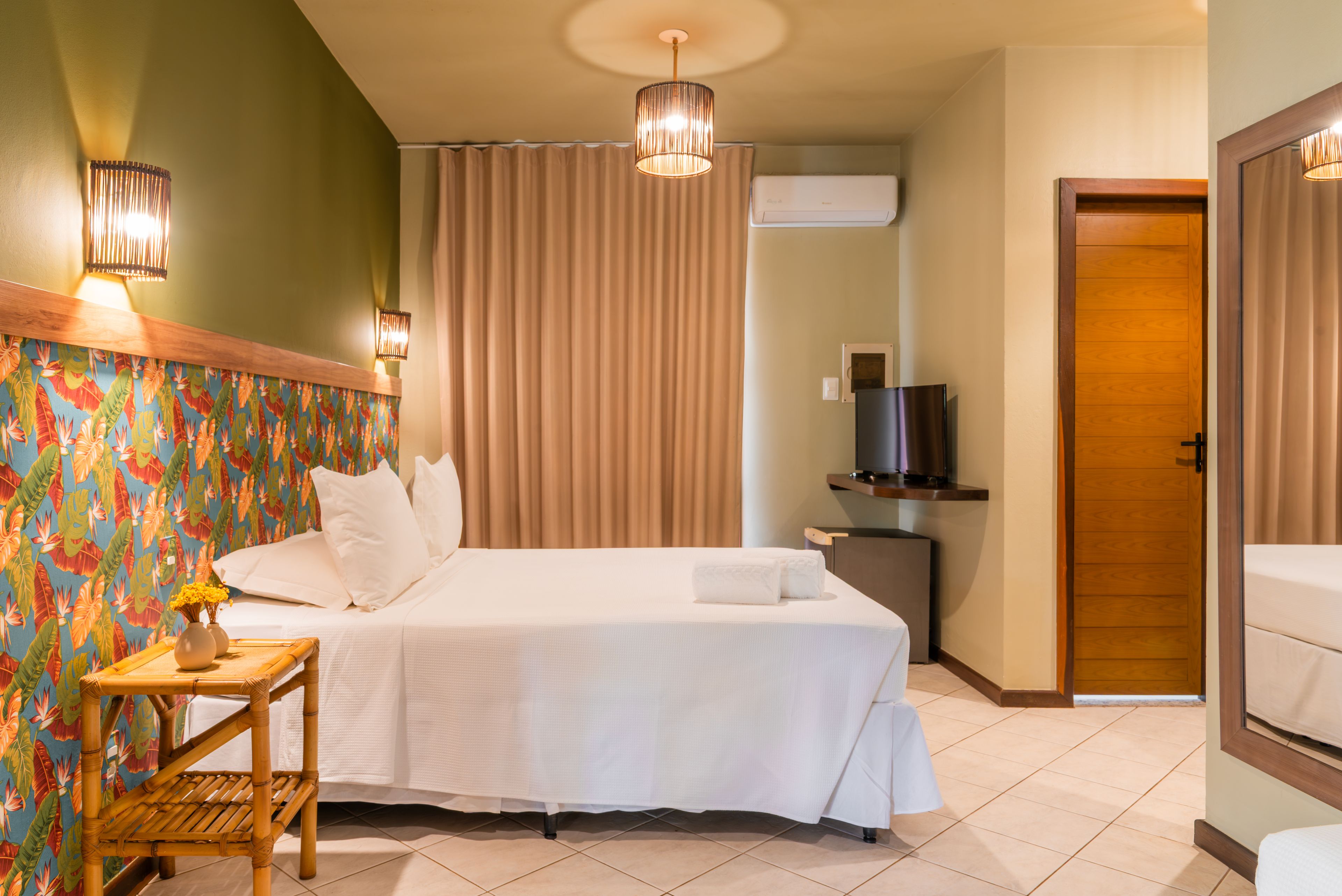 A hotel room with a bed, a television, a small table with a vase, and a wooden door.