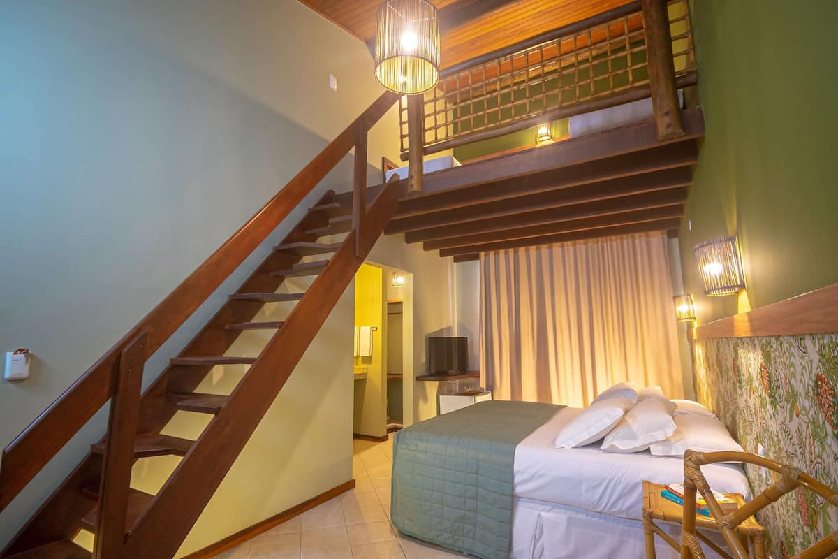 Een slaapkamer met een houten trap, een bed met witte lakens, een televisie op een standaard en een balkon met uitzicht.