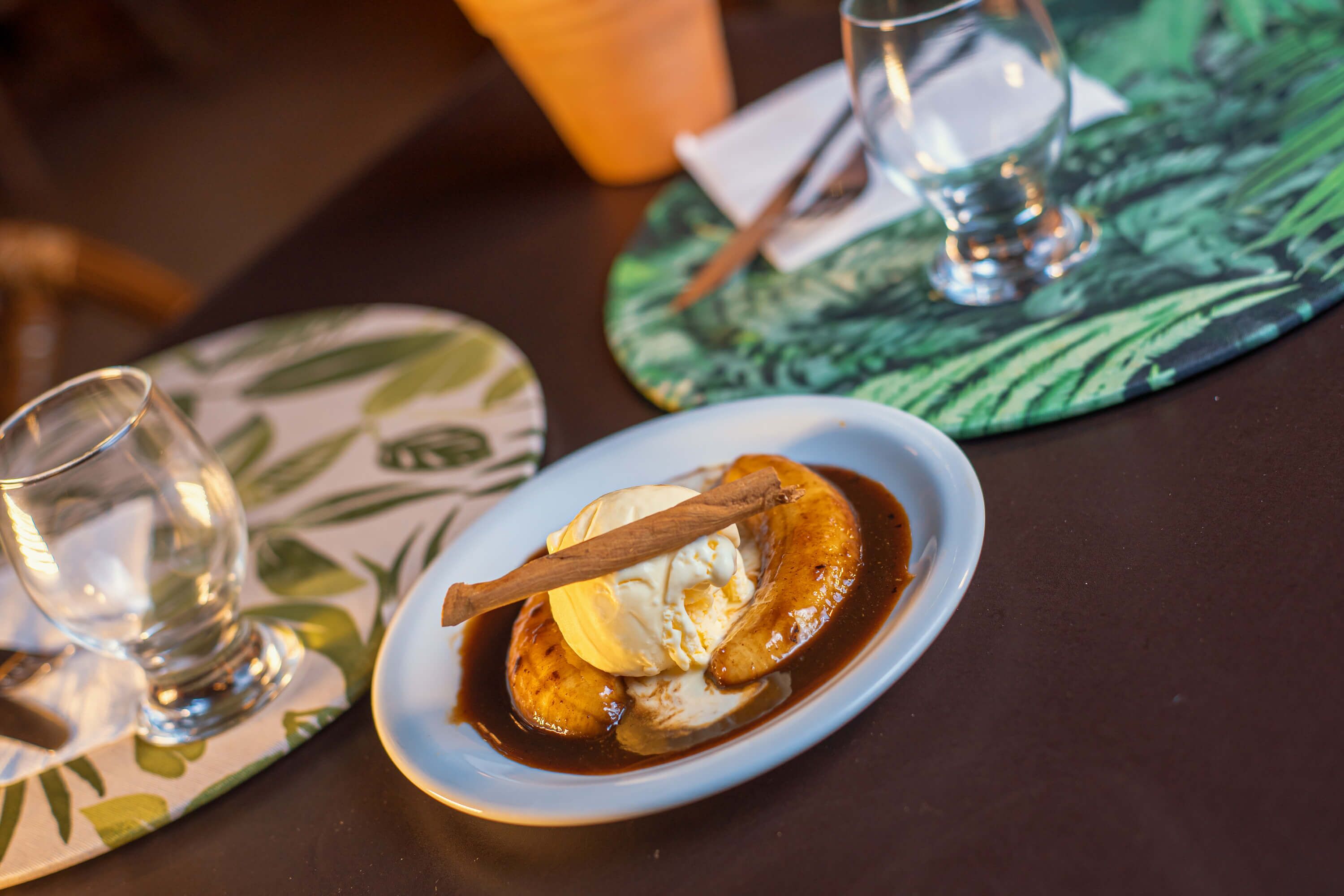 A dessert plate with a banana, ice cream, and a cinnamon stick on a table with a placemat.