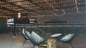 An outdoor seating area with metal chairs and a wooden table, under a roof with string lights. - Bed To Bangkok - Hostel (Bangkok)