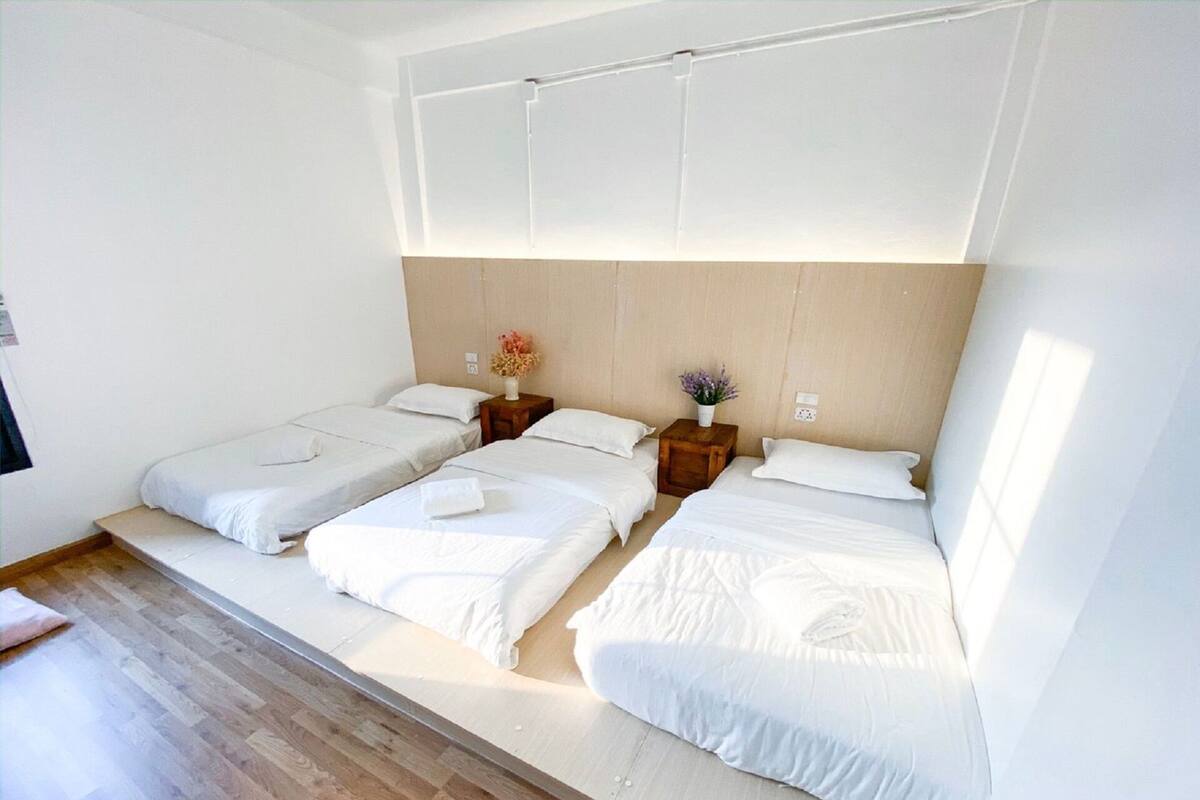 A hotel room with three single beds, wooden flooring, a wooden headboard, and a small table with a vase of flowers.