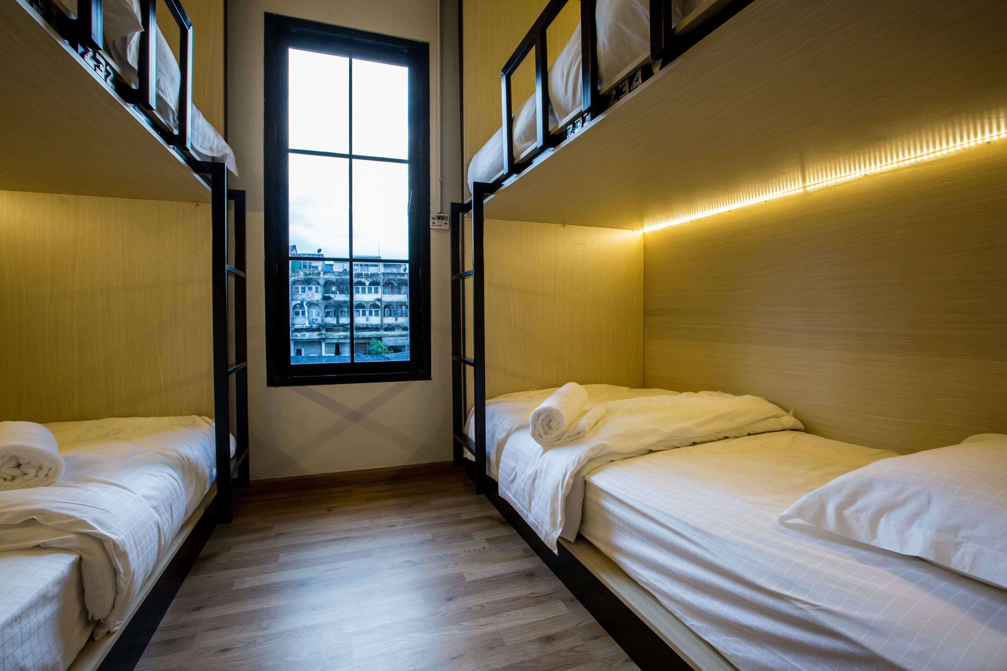 A dormitory room with bunk beds, a window with a view of buildings, and a wooden floor.