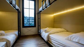 A dormitory room with bunk beds, a window with a view of buildings, and a wooden floor.