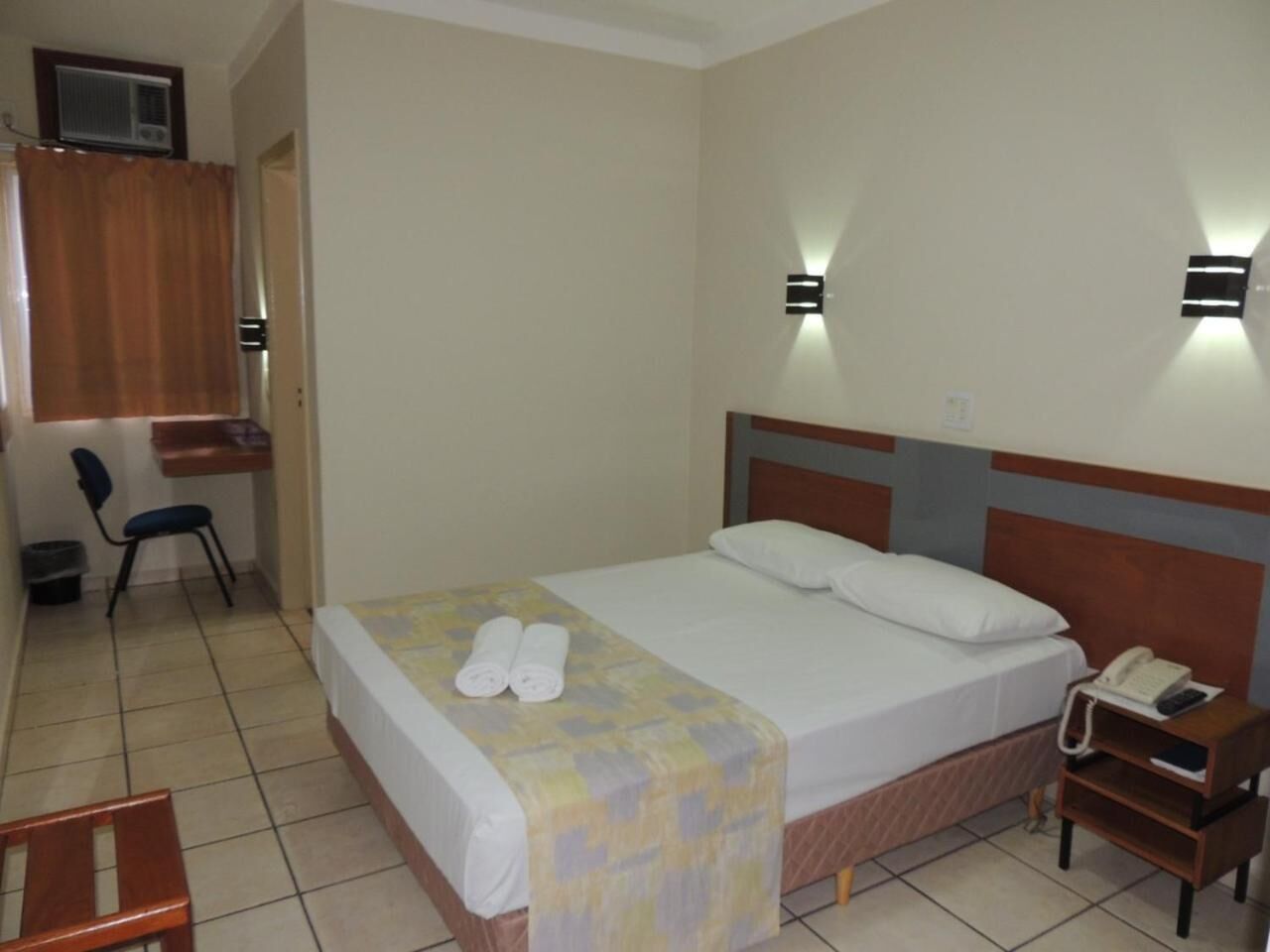 A hotel room with a single bed, a bedside table with a phone, a chair, a desk, and a wall-mounted light fixture.