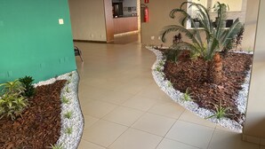 A hotel lobby with a reception desk, a fire extinguisher, and a plant area with white pebbles and mulch. - Pérola Verde Hotel (Birigui)