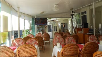 An indoor dining area with wicker chairs and tables, a television screen, and a view of the outside.