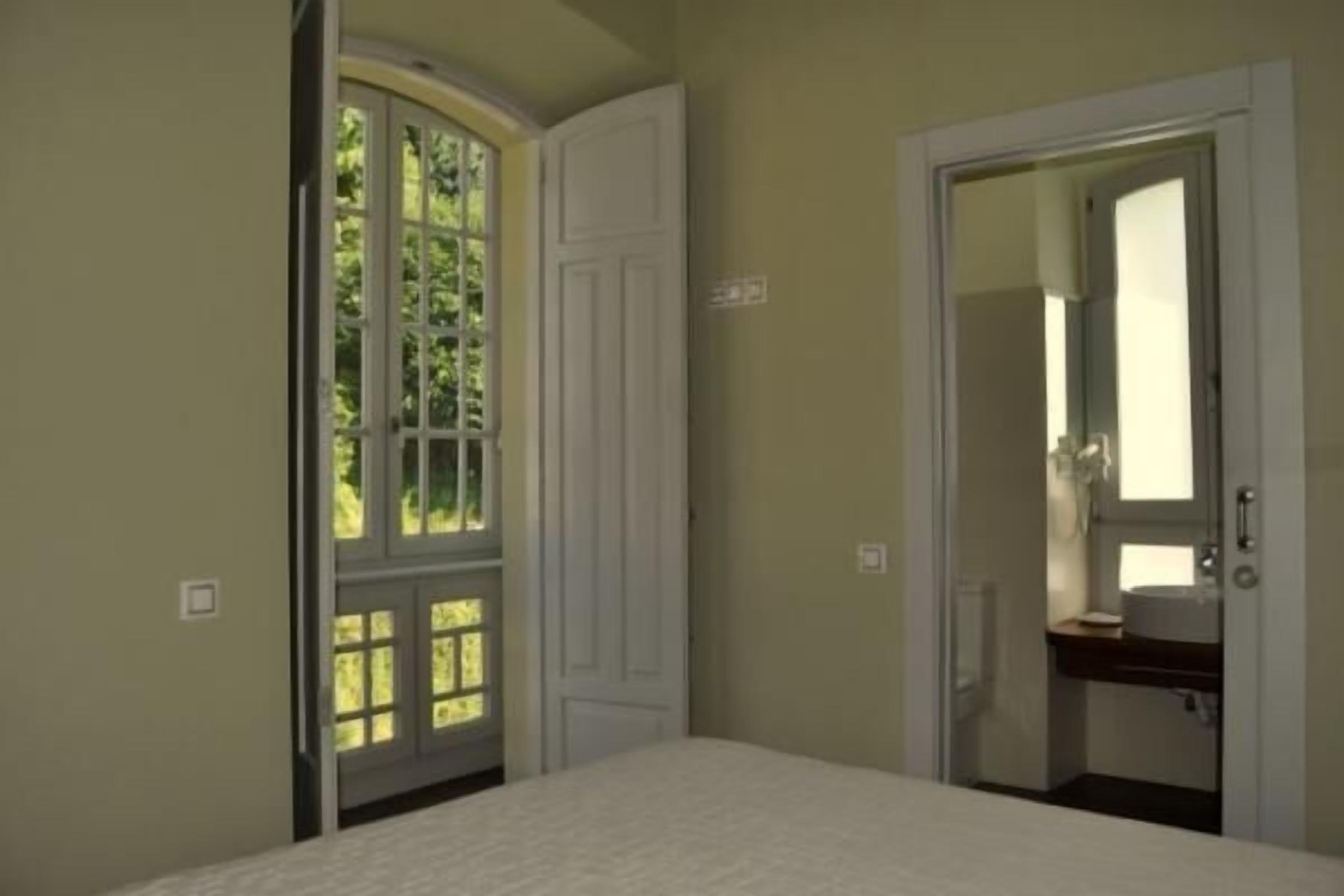 A bedroom with a bed, a door leading to a bathroom, and a window with a view of greenery.