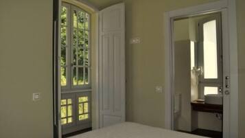 A bedroom with a bed, a door leading to a bathroom, and a window with a view of greenery.