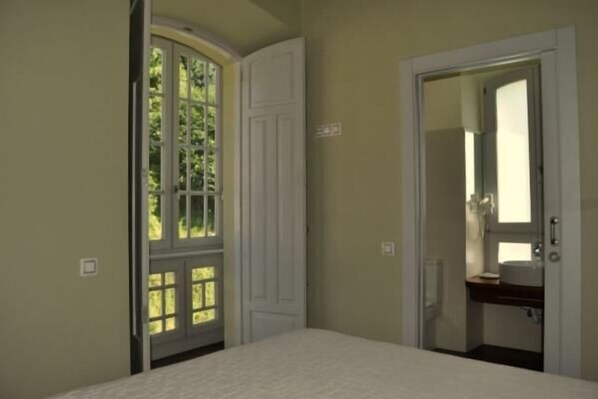 Un dormitorio con una cama, una puerta que da a un baño y una ventana con vistas a la vegetación.