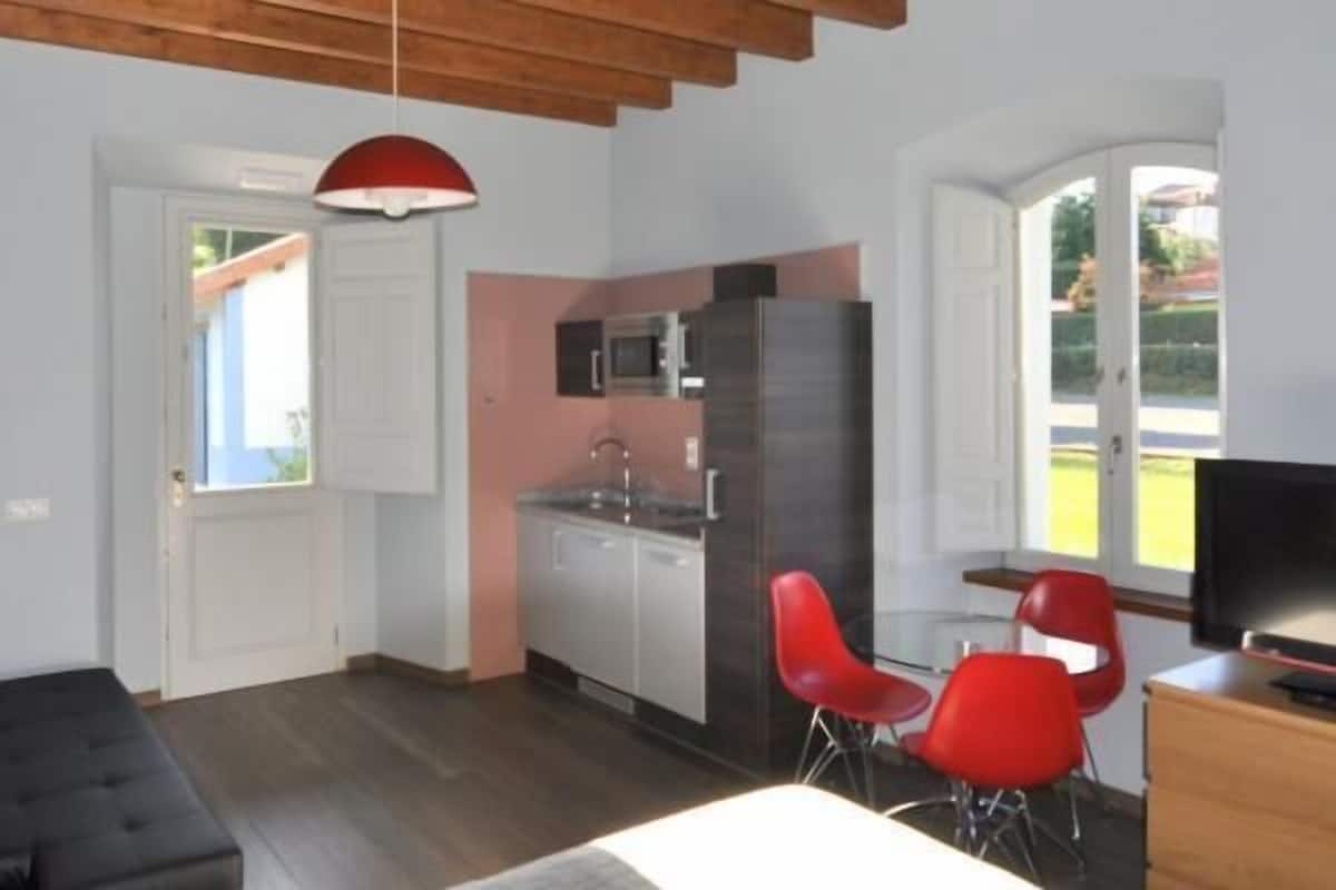 Un espace de vie compact comprenant un coin cuisine, un coin repas avec des chaises rouges, un téléviseur et une grande fenêtre.