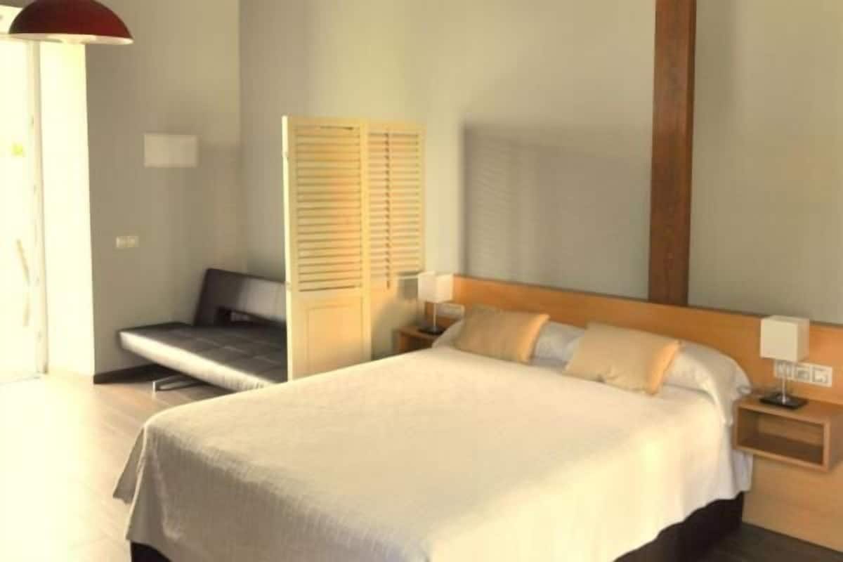 Une chambre d’hôtel comprenant un lit, des tables de chevet, une commode, un banc et une fenêtre avec des stores.