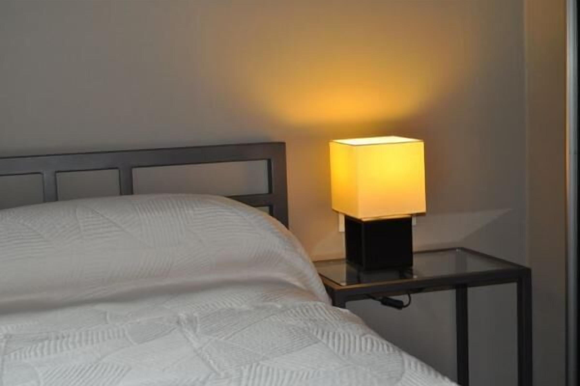 Ein ordentlich bezogenes Bett mit Kopfteil, ein Nachttisch mit Lampe und eine Wand mit strukturierter Oberfläche.