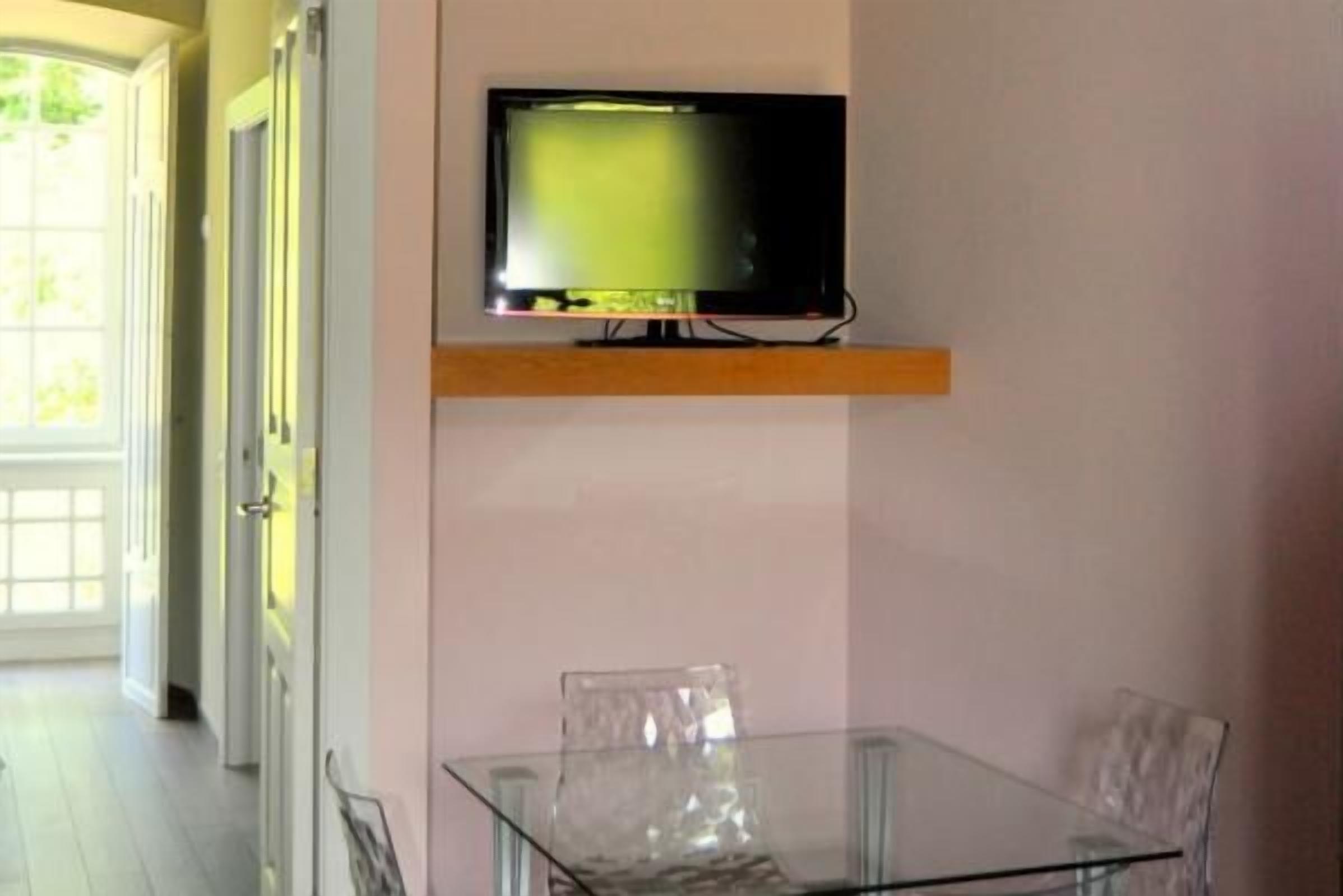 Una habitación con una mesa y sillas de vidrio, un televisor montado y un estante de madera.