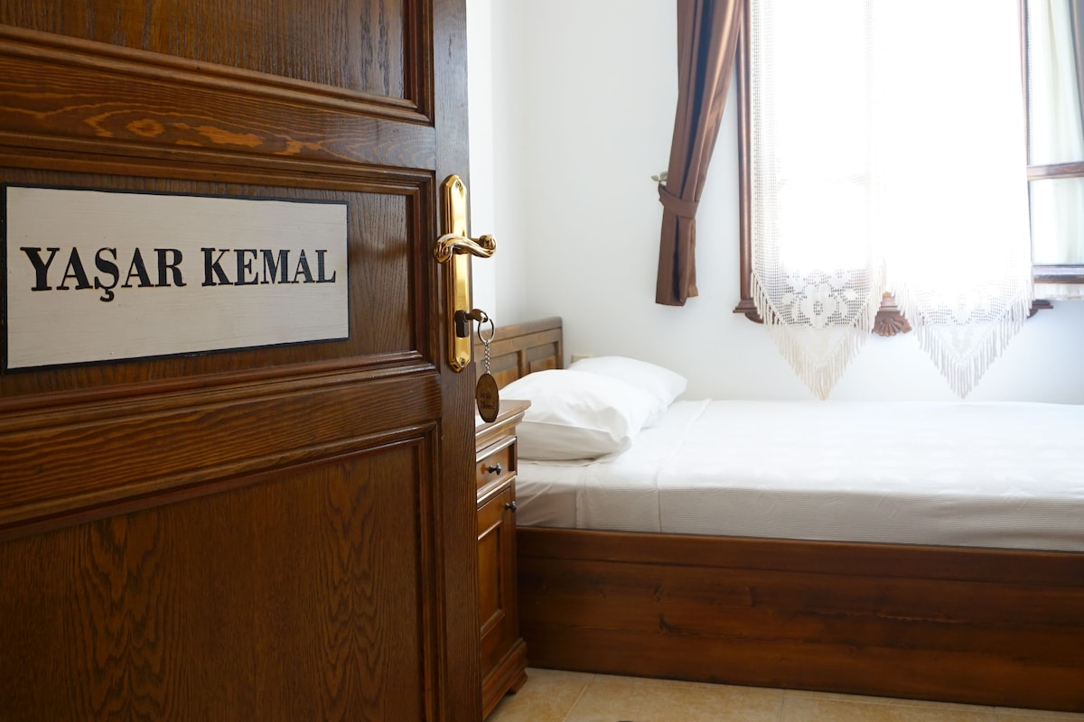 Habitación de hotel con puerta de madera, cama con ropa blanca, mesita de noche pequeña, alfombra decorativa y ventana con cortinas.