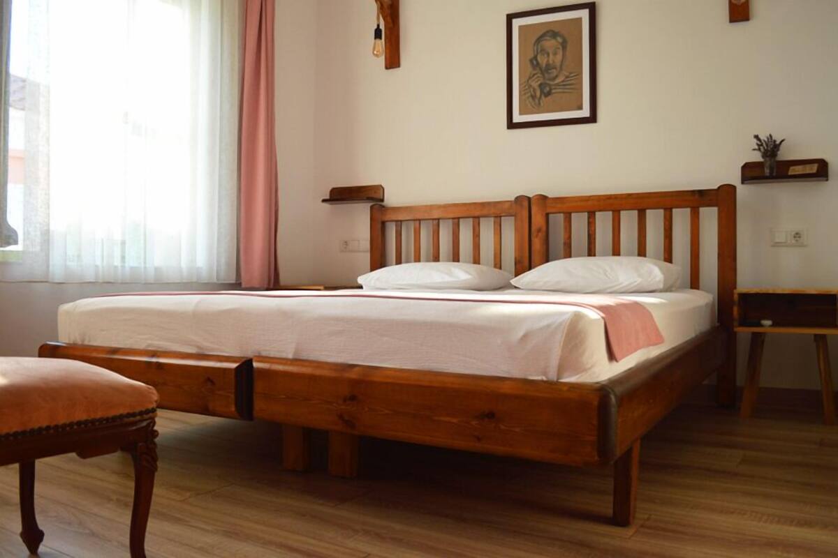 Ein hölzernes Bett mit weißer Matratze, ein Nachttisch aus Holz, ein Stuhl und ein Fenster mit transparenten Vorhängen.