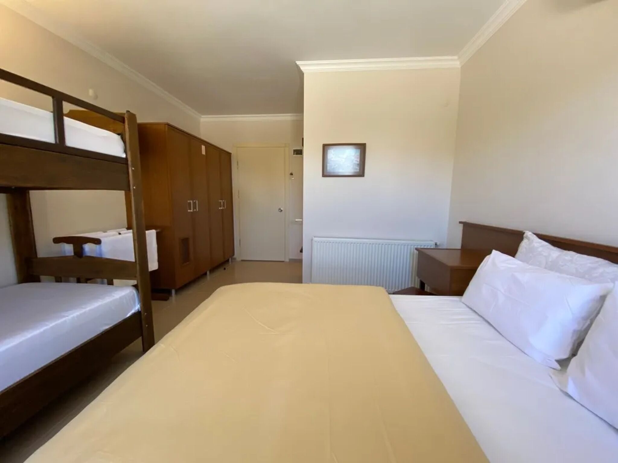 A room with a bunk bed, a single bed, wooden wardrobes, a radiator, and a framed picture on the wall.