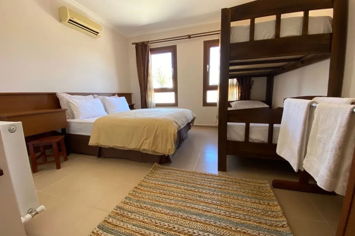 A room with a bunk bed, a single bed, a wooden bench, a small stool, and a patterned rug.