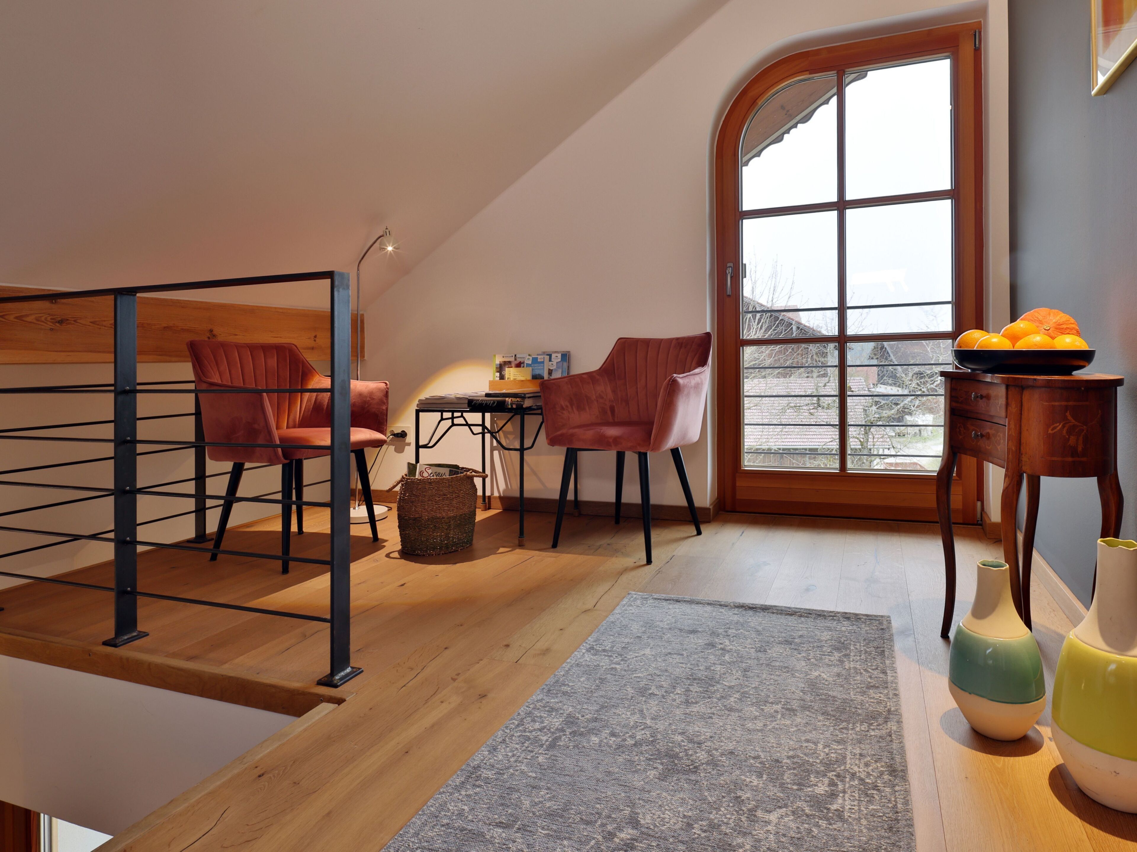 Un dormitorio tipo loft moderno con techo inclinado, piso de madera y un ventanal grande. Hay dos sillas, una mesita y un florero en el suelo.
