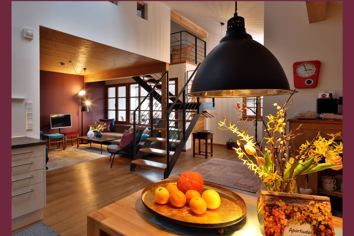 A modern living room with a wooden floor, a black pendant light, a staircase, and a dining area with a fruit bowl.