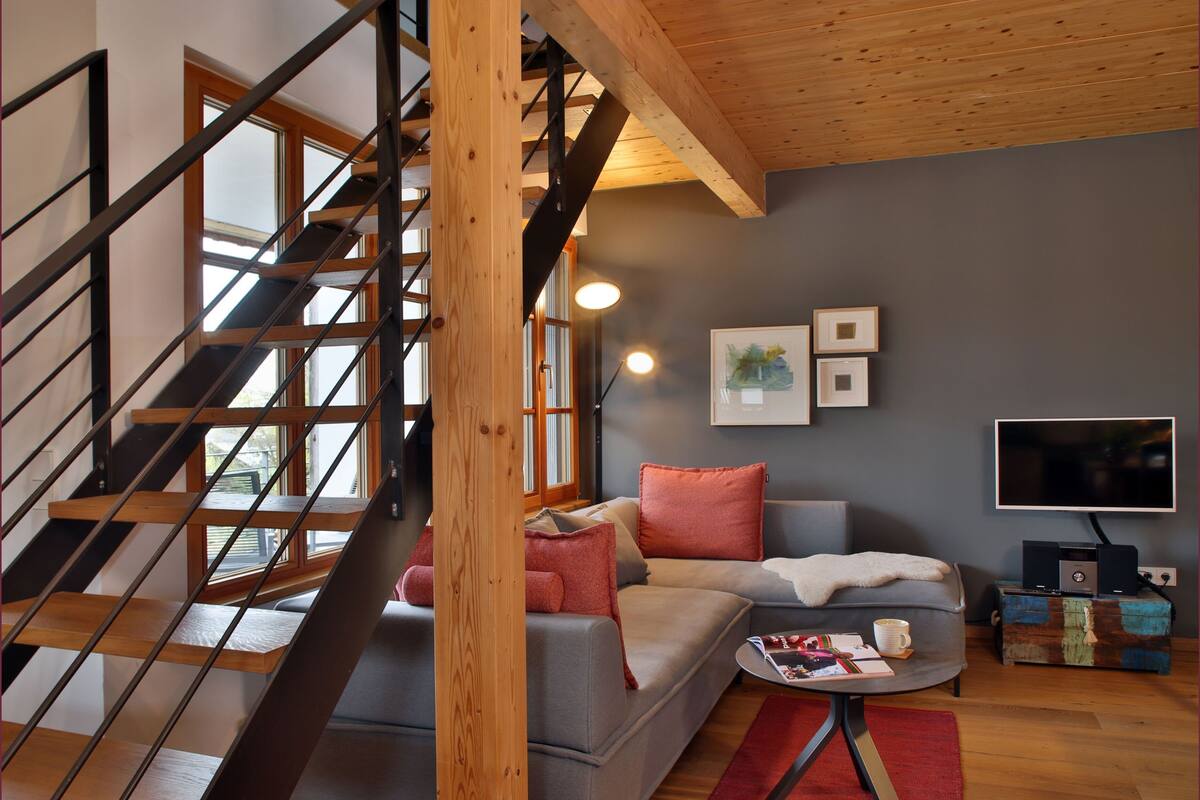A modern living room with a staircase, grey sofa, red cushions, a small table, and a TV.