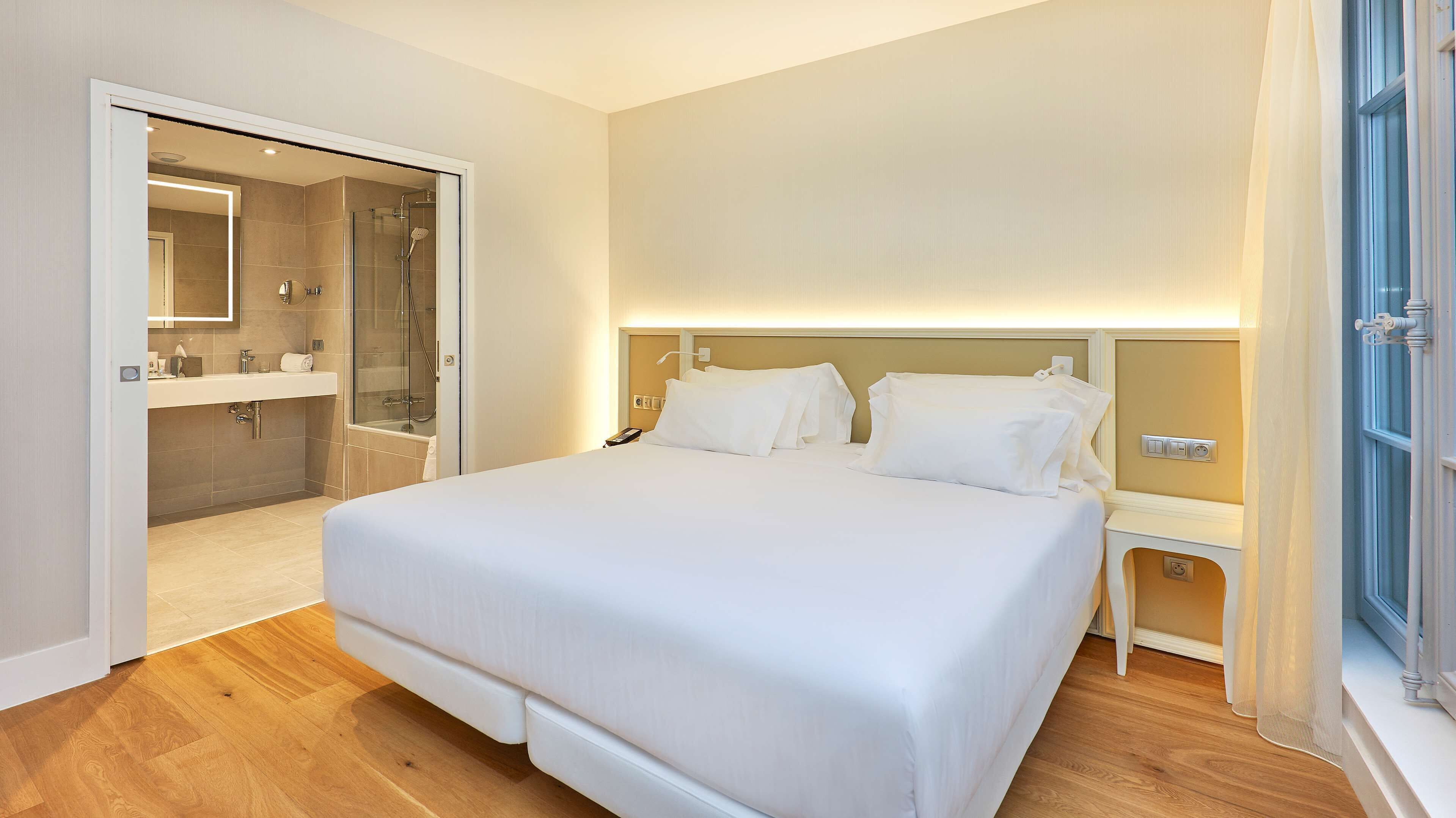 Ein modernes Hotelzimmer mit einem großen Bett, Nachttischen, einer gläsernen Duschkabine und einer neutralen Farbgestaltung.