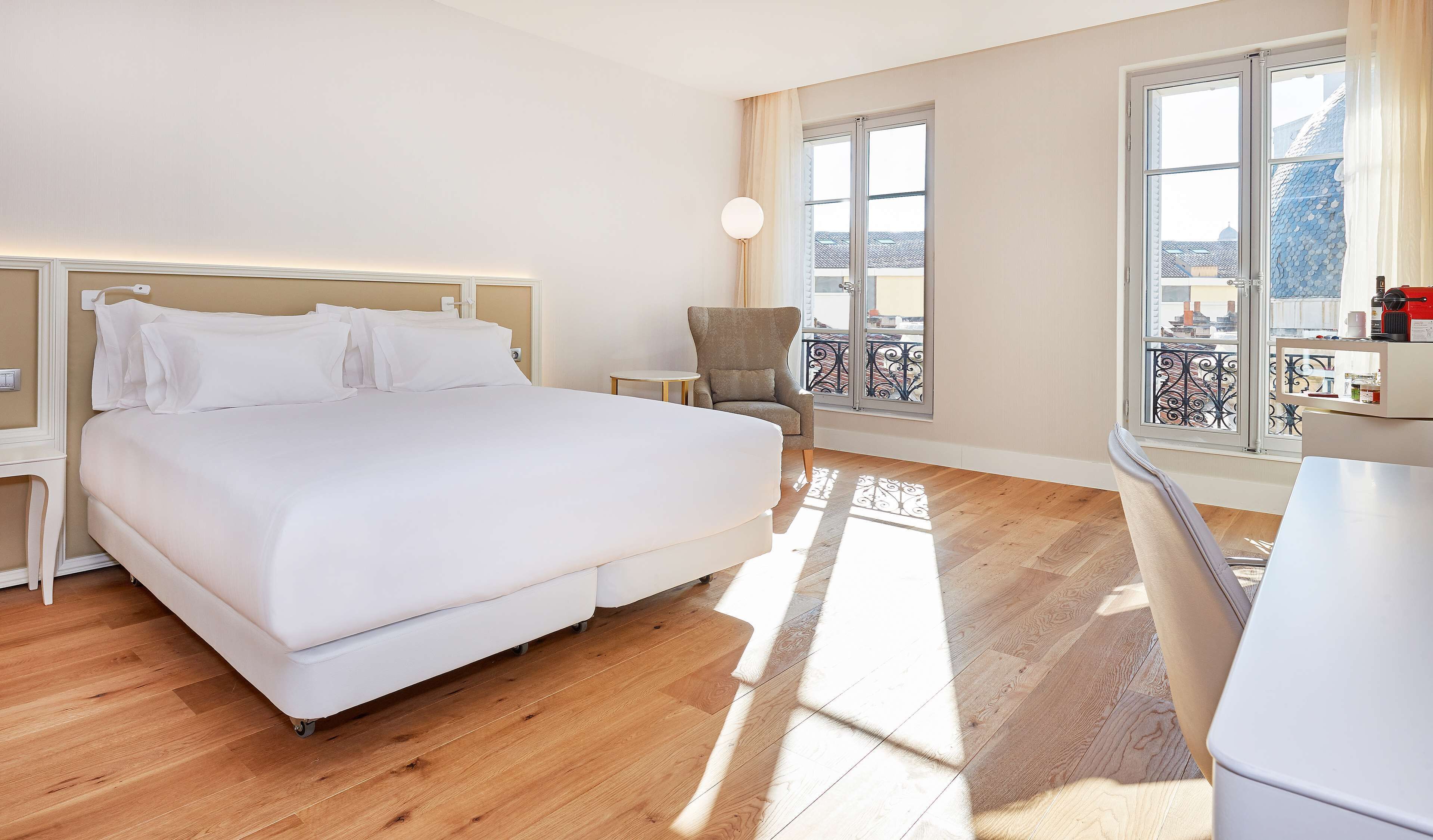 A hotel room with a large bed, a small round table, a chair, and a view of the city through large windows.