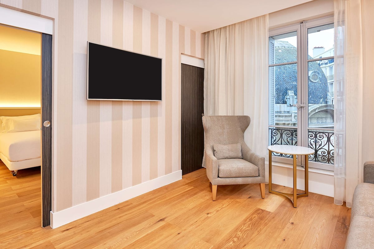 Une chambre d’hôtel avec un lit, un téléviseur fixé au mur, une chaise, une petite table et une fenêtre donnant sur des immeubles.