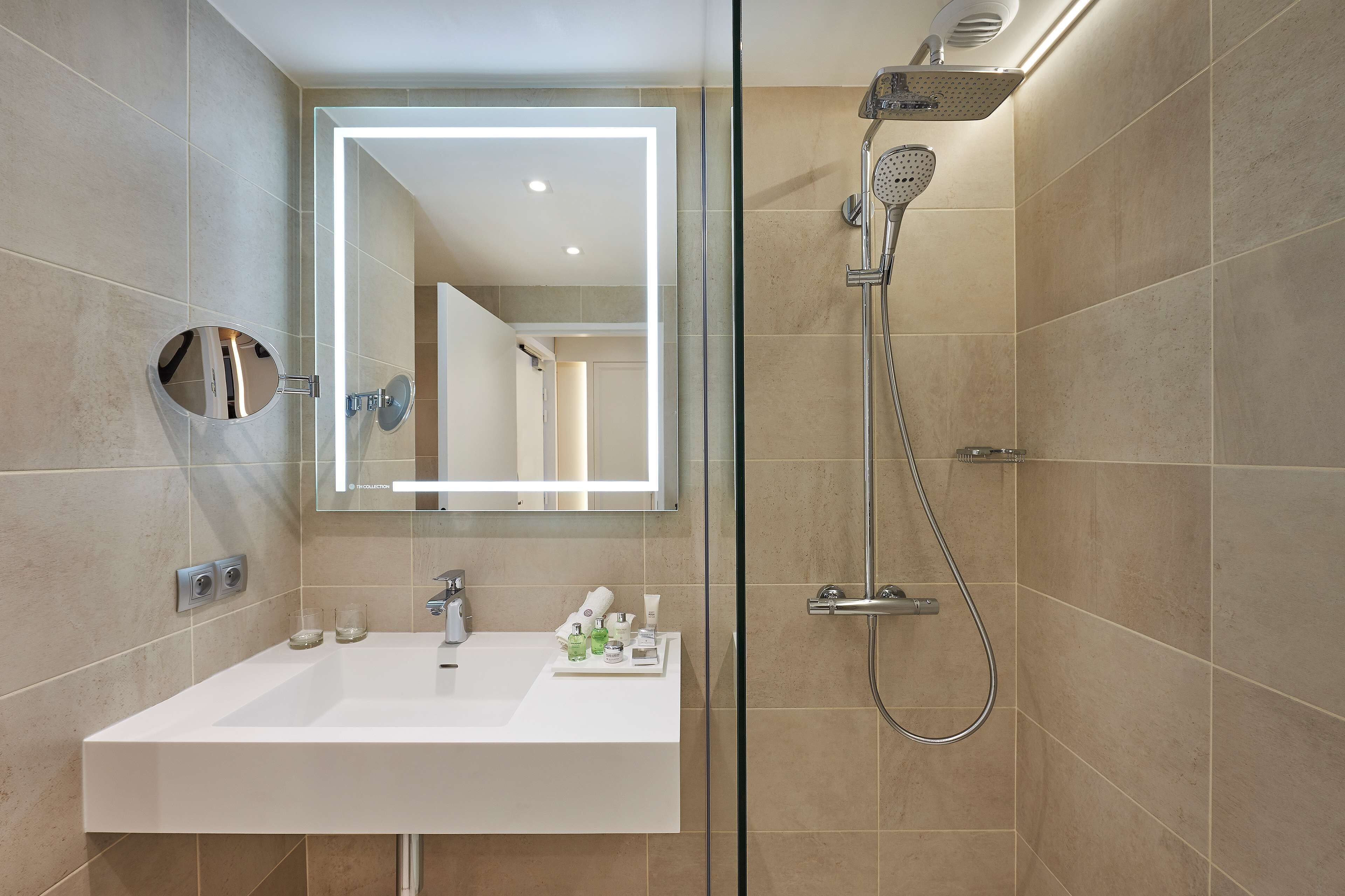 A modern bathroom with a white sink, a mirror with integrated lighting, a handheld showerhead, and a glass-enclosed shower.